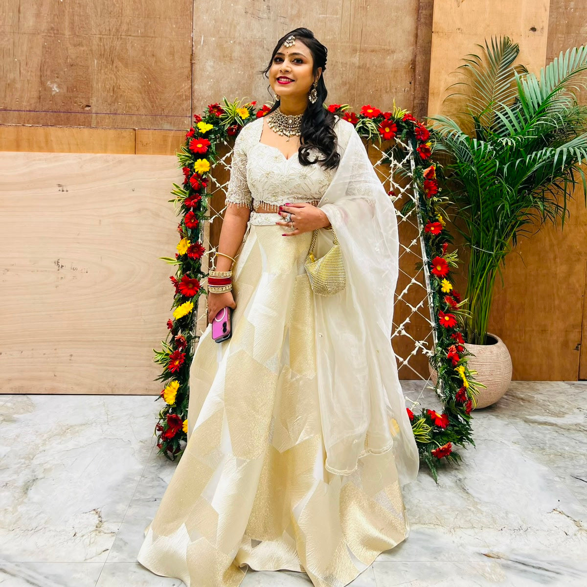 Woman in a white traditional outfit standing in front of a decorated entrance with flowers and plants.