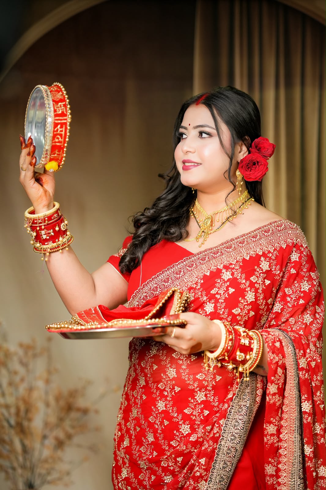 A woman in a red embroidered traditional outfit holds a mirror and a plate with a red shoe, set against a warm indoor backdrop with curtains.
