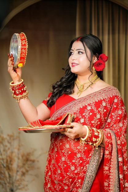 A woman in a red embroidered traditional outfit holds a mirror and a plate with a red shoe, set against a warm indoor backdrop with curtains.