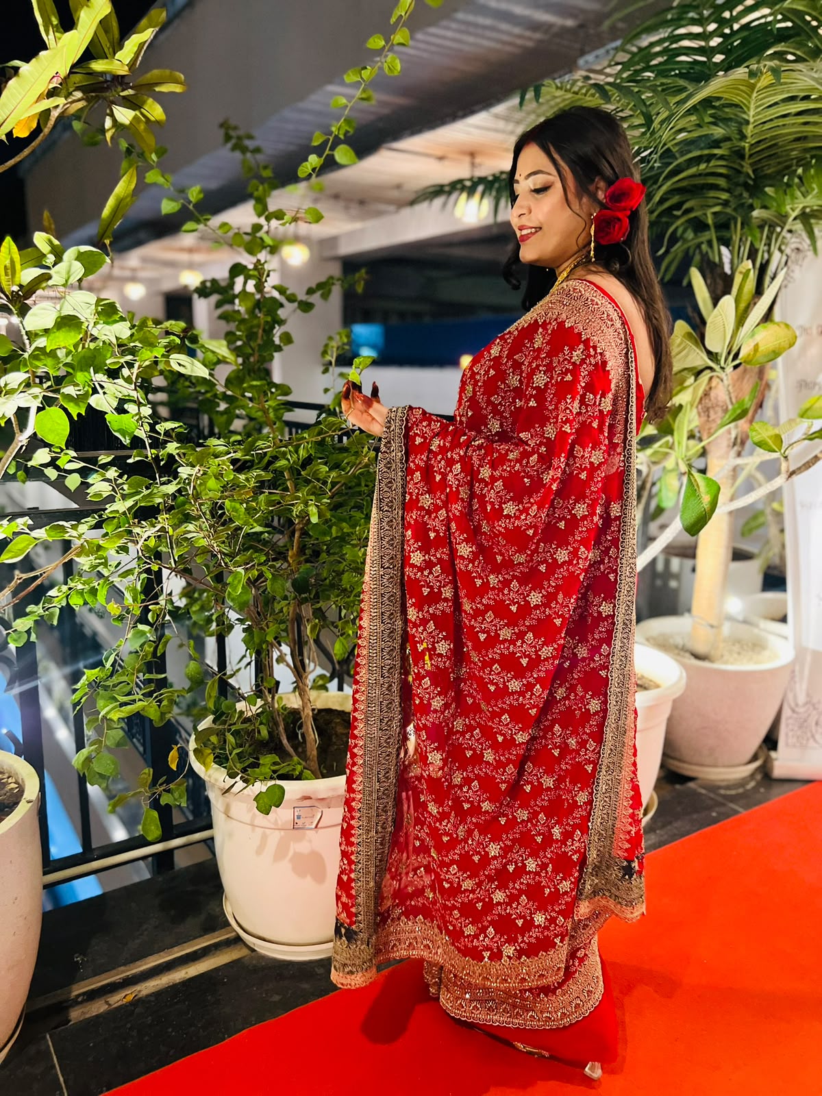 A woman wearing a red embroidered traditional dress with a floral pattern and a matching dupatta, standing on a red carpet in front of potted plants.