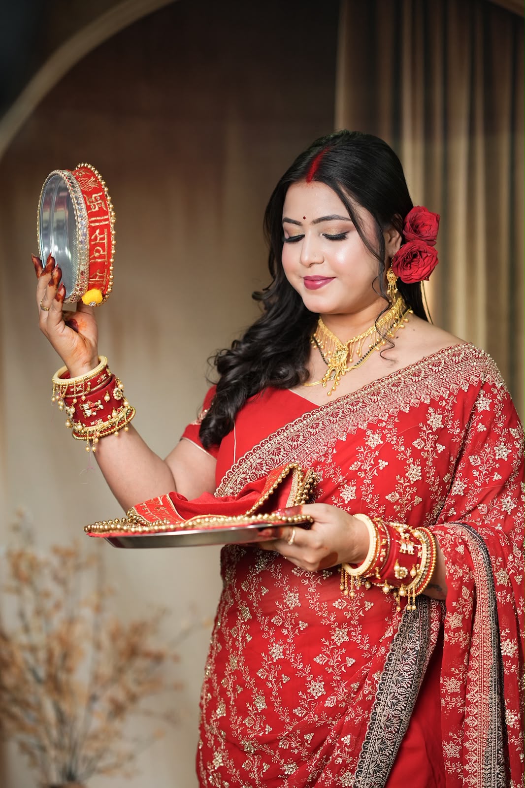 A woman in a red and gold embroidered traditional sari holds a silver mirror and a decorative tray during what appears to be a ceremonial ritual. She is adorned with red flowers in her hair, a red bindi, and multiple gold and red bangles and necklaces.