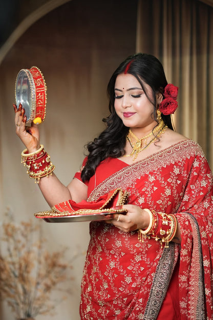A woman in a red and gold embroidered traditional sari holds a silver mirror and a decorative tray during what appears to be a ceremonial ritual. She is adorned with red flowers in her hair, a red bindi, and multiple gold and red bangles and necklaces.