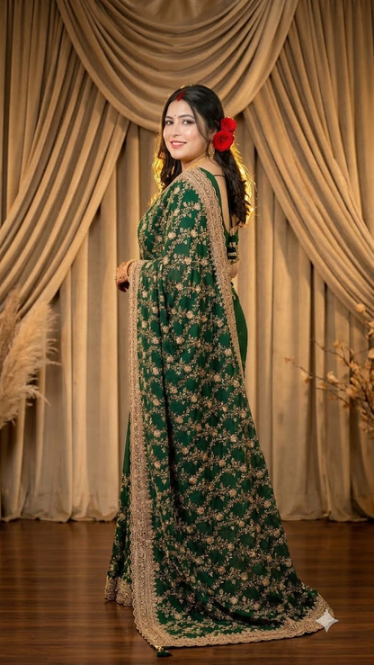 A woman wearing a dark green embroidered saree with gold floral patterns and a beige border, standing in front of draped beige curtains.