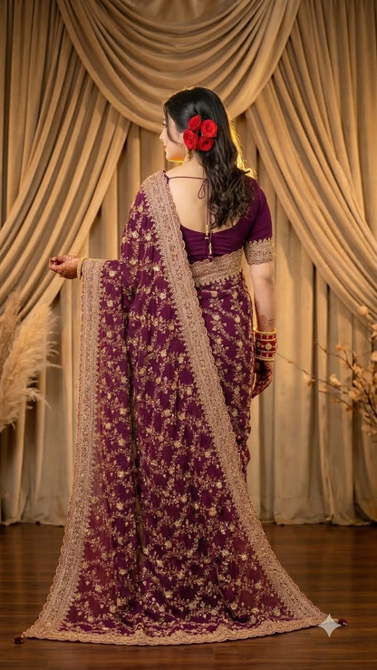 A woman stands with her back to the camera wearing a deep purple embroidered saree with a beige border and a matching purple blouse, posed in front of draped beige curtains.