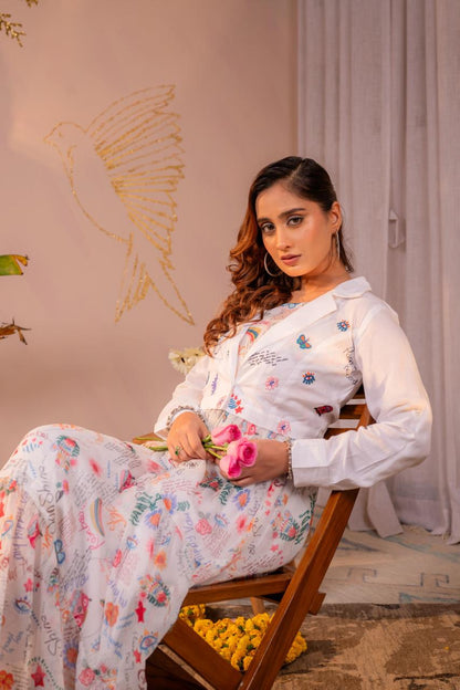 Seated close-up of woman wearing white doodle print maxi dress with embroidered cropped blazer holding pink roses - Jagranee