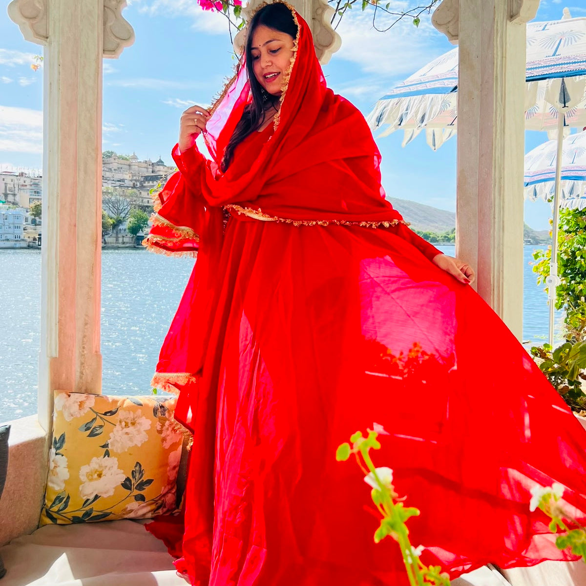 Red georgette Anarkali gown with dupatta draped over head showing gold lace trim, white marble palace with lake backdrop - Jagranee