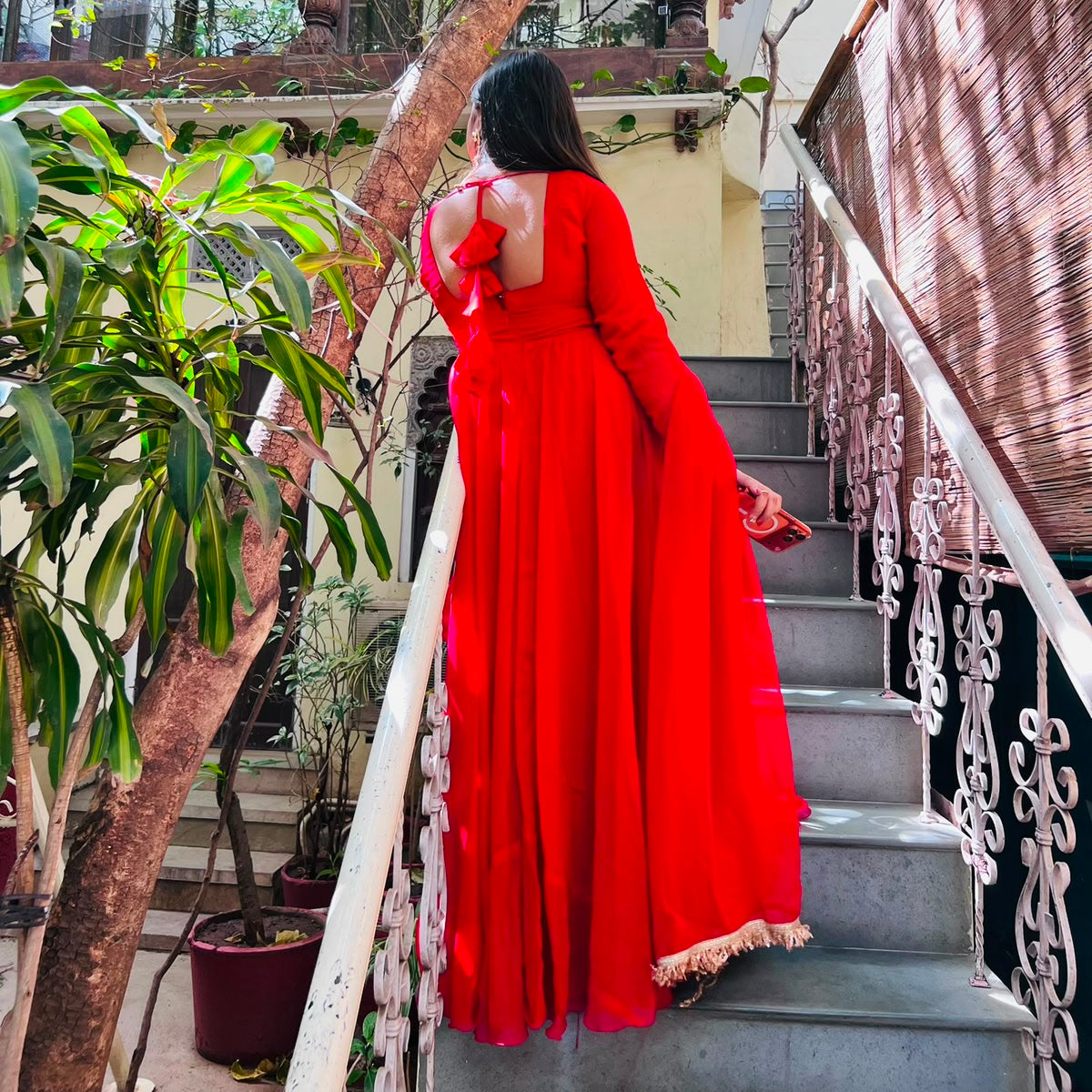 Back view of red georgette Anarkali gown showing tie-back bow detail and gold lace hemline on heritage haveli staircase - Jagranee